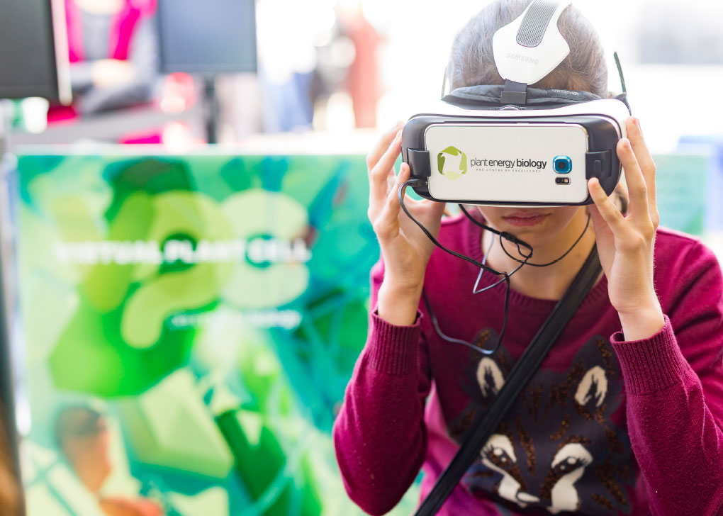 A young girl wearing red jumper, with viewer goggles strapped over her eyes watching a virtual reality video