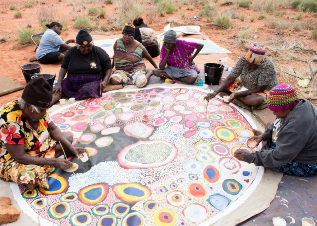 Several Aboriginal women sitting on sandy red earth, working together on one large circular painting that features many brightly coloured circular designs.