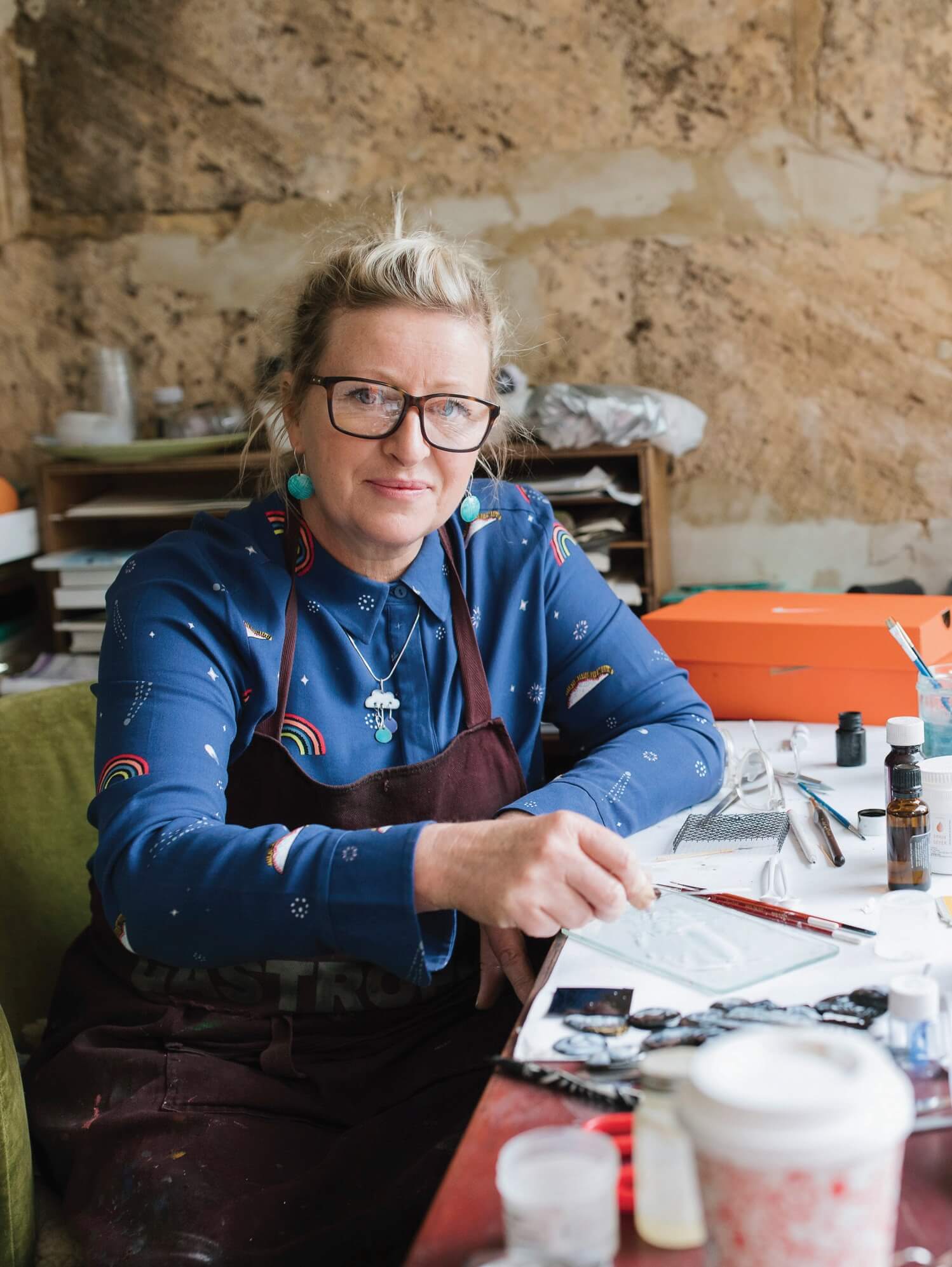 Woman with blonde hair, wearing black glasses and a maroon apron, seated on a green chair at a desk covered with craft-making materials.