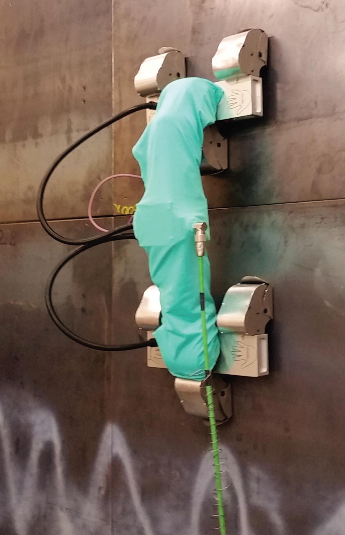 A green autonomous climbing robot climbing up a grey, glossy metallic wall. 