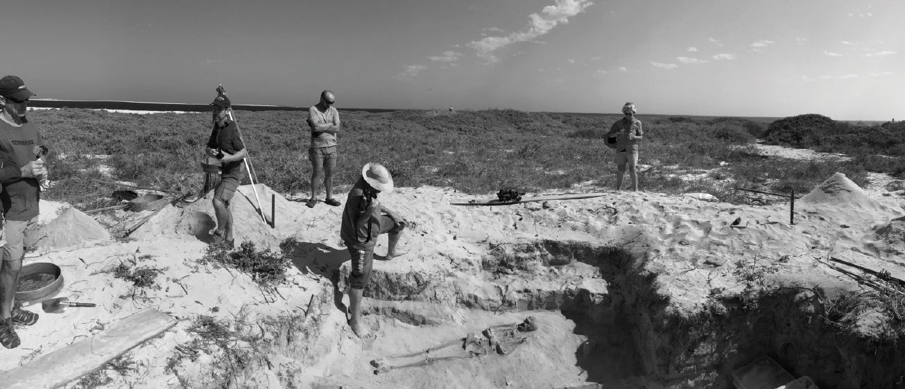 Black and white photograph of researchers gathered around dig revealing skeleton.